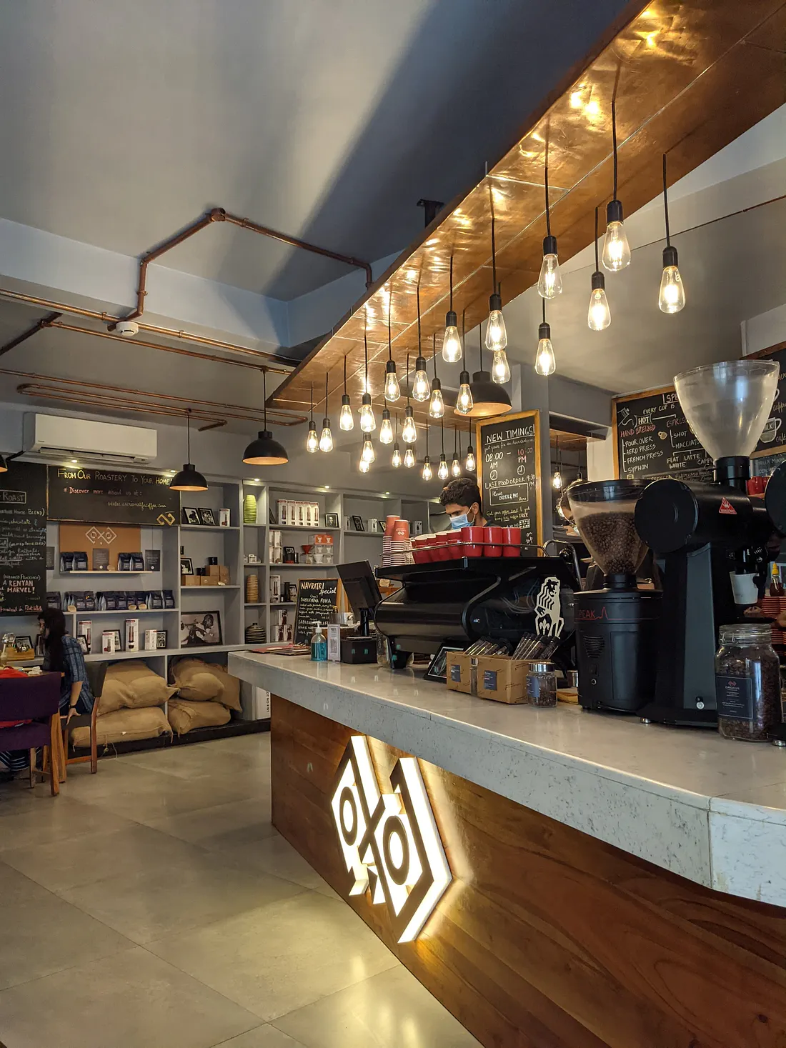 The service counter of a Jaipur specialty coffee roastery, lit by a row of hanging Edison bulbs, with retail bean shelves behind it
