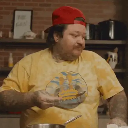 A chef pulling a loaded spoon back out of his mouth mid-taste, eyes shut, pan still in hand